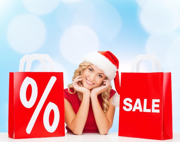 verkoop, geschenken, Kerstmis, vakantie en mensen concept - lachende vrouw in santa helper hoed met boodschappentassen en procentteken over blauwe lichten achtergrond