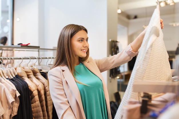 verkoop, winkelen, mode, stijl en mensenconcept - gelukkige jonge vrouw die kleding kiest in winkelcentrum of kledingwinkel