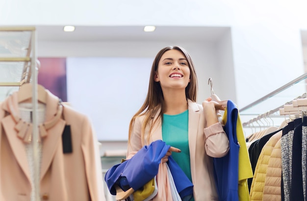verkoop, winkelen, mode, stijl en mensenconcept - gelukkige jonge vrouw die kleding kiest in winkelcentrum of kledingwinkel