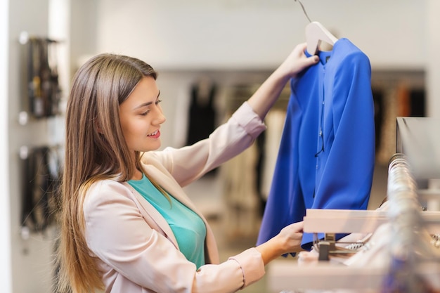 verkoop, winkelen, mode, stijl en mensenconcept - gelukkige jonge vrouw die kleding kiest in winkelcentrum of kledingwinkel