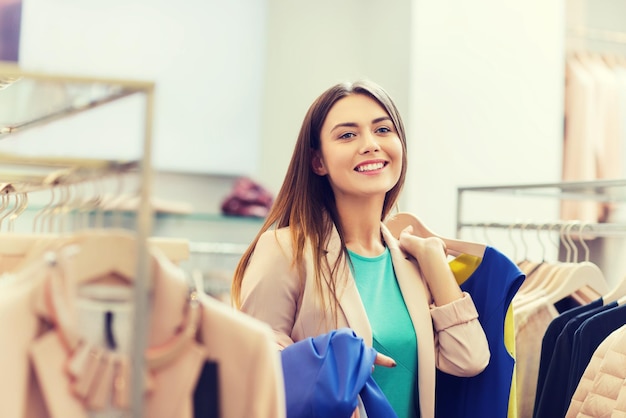 verkoop, winkelen, mode, stijl en mensenconcept - gelukkige jonge vrouw die kleding kiest in winkelcentrum of kledingwinkel