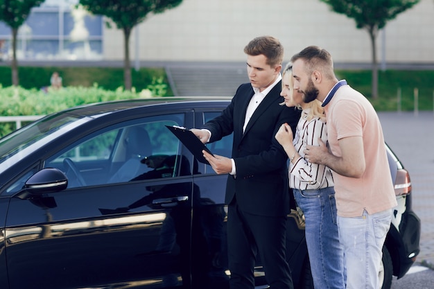 Foto verkoper en een jong stel buiten in de buurt van een nieuwe auto. de verkoper vertelt het jonge stel over de auto. man en vrouw kopen een auto.