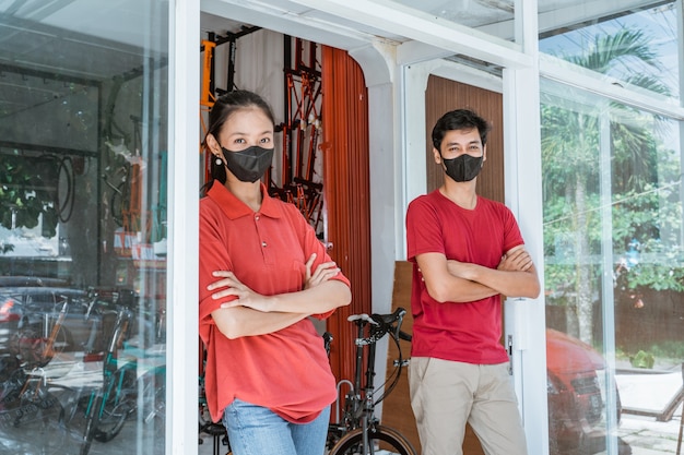 Verkoper en verkoopster met masker staan met gevouwen handen voor de glazen deur te leunen