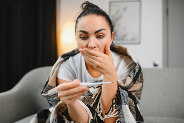 Verkoudheid en griep Zieke vrouw betrapt koude gevoel ziekte Close-up van mooi ongezond meisje bedekt met deken neus afvegen en kijken naar thermometer Gezondheidszorg concept