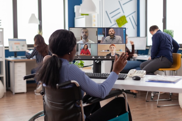 Verlamde gehandicapte zwarte werknemer zittend geïmmobiliseerd in rolstoel met videovergadering online bespreken met collega's op afstand in kantoor business