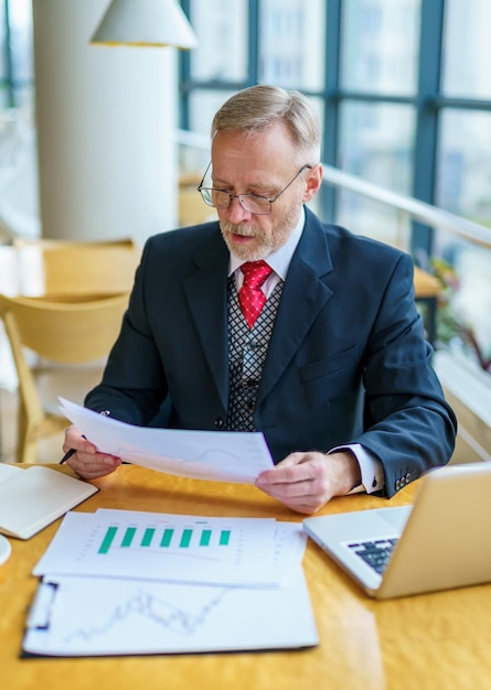 Verlaten zakenman die op zijn papierwerk kijkt Volwassen man in stijlvol pak doet zijn werk met laptop in gezellig café