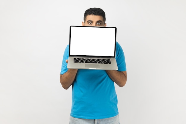 Verlegen jonge volwassen man die de helft van zijn gezicht verbergt achter een laptop met een leeg scherm dat naar de camera kijkt