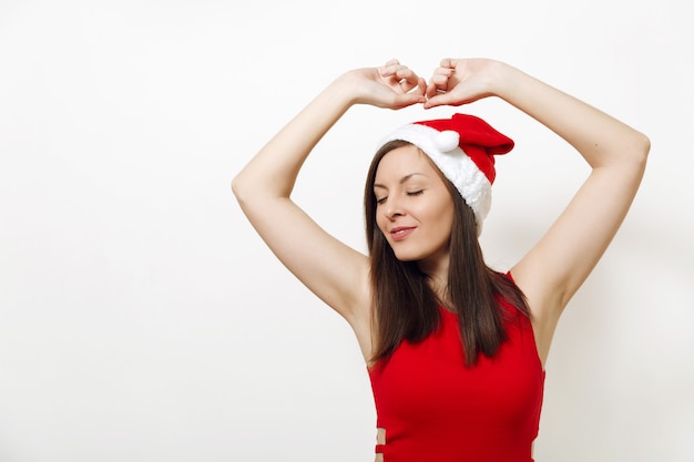 Verleidelijke blanke jonge gelukkige vrouw met ogen gesloten met plezier en charmante glimlach met rode jurk en kerstmuts op witte achtergrond. Santa meisje geïsoleerd. Nieuwjaarsvakantie 2018 concept.