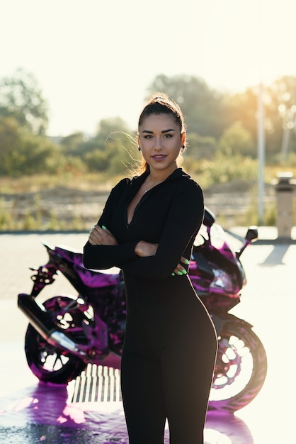 Verleidelijke jonge vrouw in strak zwart pak vormt in de buurt van sport motorfiets bij self service car wash