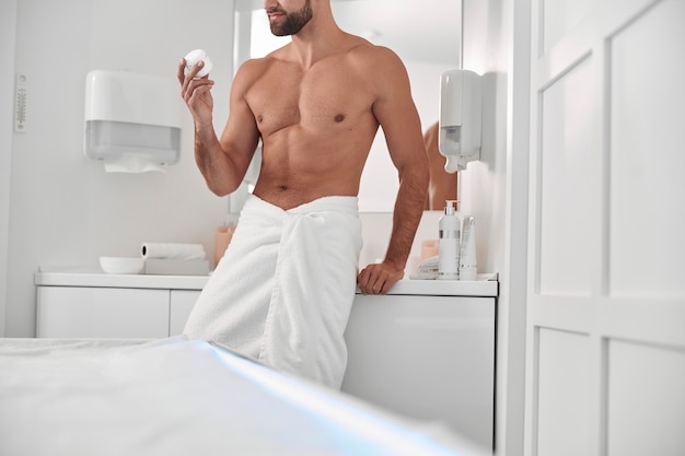 Verleidelijke man met blote borst houdt een pot crème vast die op een kast in de buurt van de spiegel leunt in een moderne, lichte badkamerclose-up