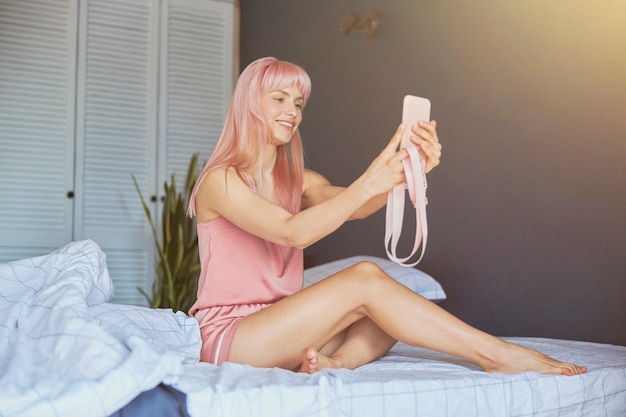 Verleidelijke vrouw met roze haar in elegante zijden pyjama neemt selfie op groot bed