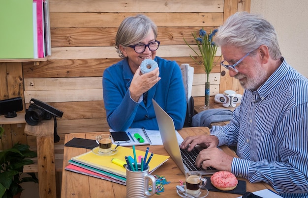 Foto verleiding donut voor een senior aantrekkelijke vrouw gepensioneerd echtpaar met wit haar zittend buiten aan een houten tafel werken met laptop en boeken