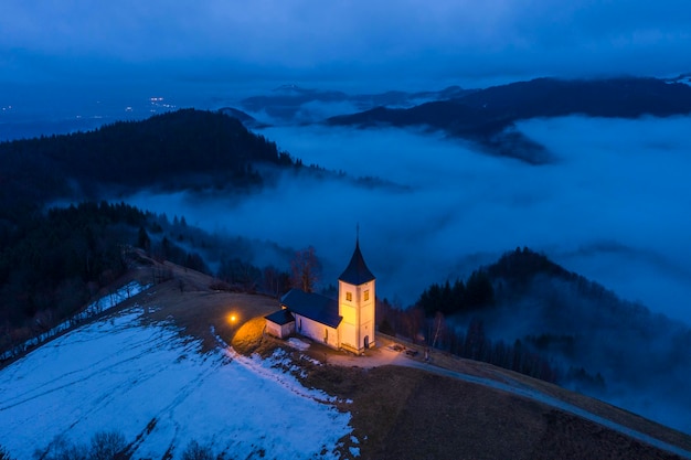 Verlichte kerk van St Primoz en Felicijan op winteravond mistige schemering bewolkte hemel Jamnik Slovenië luchtfoto