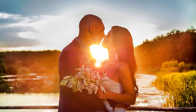 Foto verliefde paar, bruid en bruidegom kijken elkaar bij zonsondergang op een houten brug in de buurt van de rivier