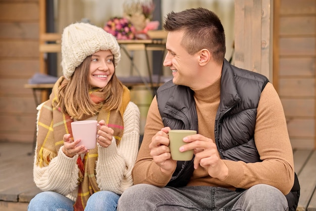 Verliefde paar met koffiemokken in hun handen zittend op de veranda en glimlachend naar elkaar