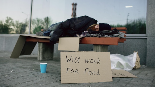 Foto verloren jonge dakloze man met cardbaord teken slaap op bankje in city street