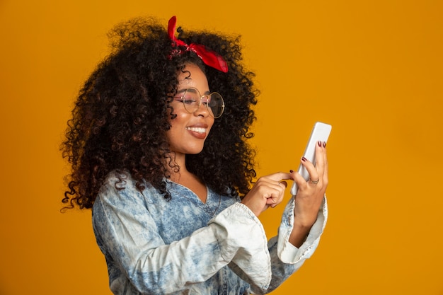 vermaakte schattig gelukkig Afro-Amerikaanse meisje met afro kapsel bedrijf smartphone met apparaat om plezier te hebben. gele muur.