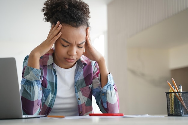 Foto vermoeid afro-amerikaans tienermeisje heeft hoofdpijn en doet huiswerk op laptop moeilijke e-learning