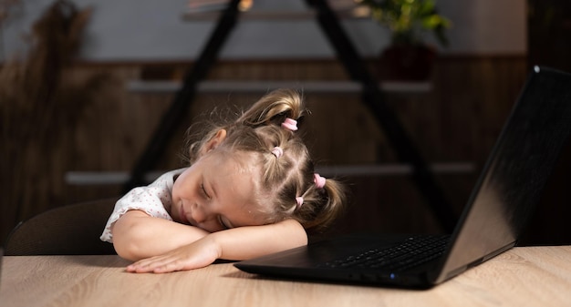 Vermoeid kleutermeisje zit aan tafel en slaapt in de buurt van de laptop