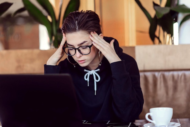Vermoeid snelkoppeling hipster meisje houdt haar hoofd vast tijdens het studeren met laptop in het café-interieur
