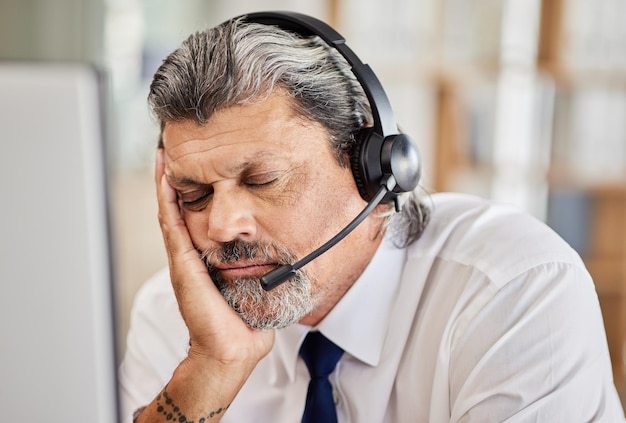 Vermoeide callcenter en mannelijke adviseur slapen in zijn kantoor tijdens een online consultatie Uitgeputte burn-out en volwassen man telemarketing of klantenservicemedewerker met een headset op de werkplek