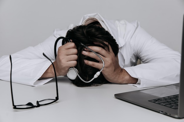 Vermoeide depressieve mannelijke arts ligt in het medische kantoor met een stethoscoop op tafel Burnout-concept