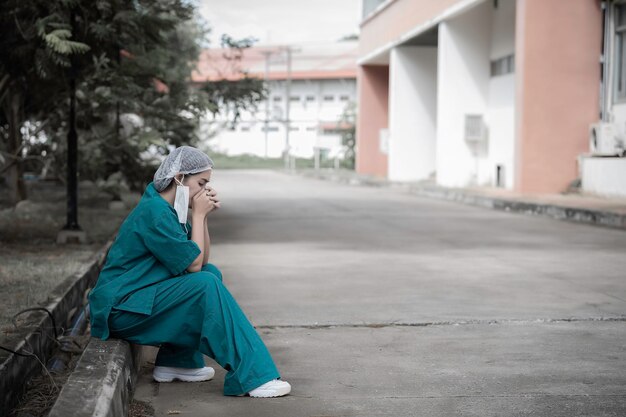 Vermoeide depressieve vrouwelijke Aziatische scrubverpleegster draagt gezichtsmasker blauw uniform zit op ziekenhuisvloer Jonge vrouwelijke arts gestrest door hard werken