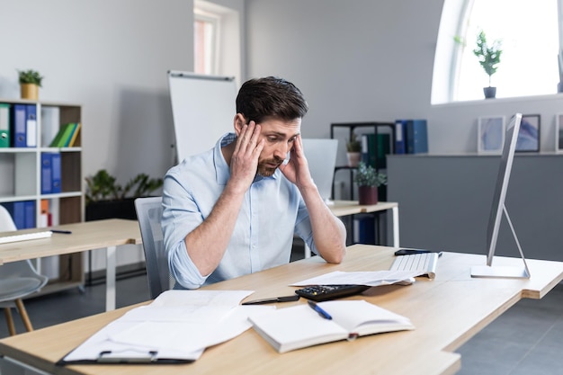 Vermoeide en geërgerde jonge man kantoormedewerker manager die zijn hoofd vasthoudt, pijn voelt duizeligheid Zittend aan een computerbureau op kantoor