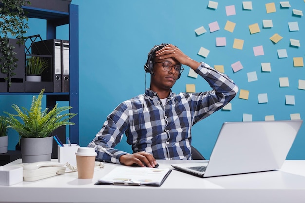 Vermoeide en gestresste klantondersteuningsoperator met hoofdpijn in telegesprek met potentiële klant. Vermoeide adviseur agent werknemer met burn-out syndroom zit aan bureau in kantoor werkruimte.