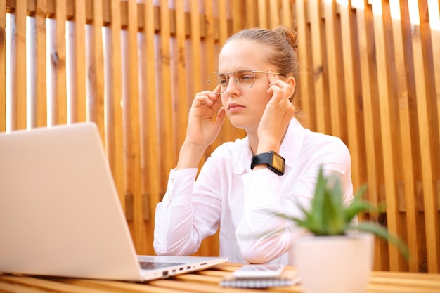 Vermoeide geconcentreerde vrouw met een wit overhemd zittend op een terras terwijl ze aan een laptop werkt en tempels masseert, heeft hoofdpijn bij het nadenken over nieuwe taken