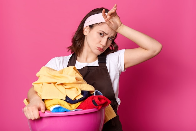 Vermoeide, hardwerkende vrouw met fronsend gezicht die haar voorhoofd met de hand aanraakt, zweet wegveegt, veel huishoudelijk werk heeft, schone kleren uit de was haalt, verdrietig kijkt.