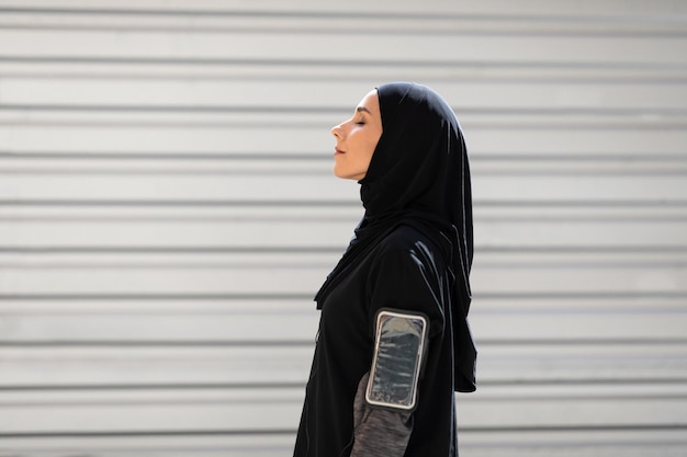 Vermoeide jonge Arabische vrouw in hijab met telefoon op schouder rustend na oefening op grijze muur