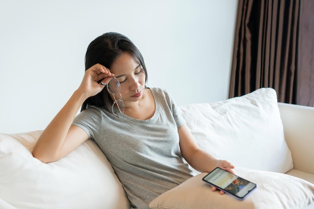 Vermoeide jonge Aziatische vrouw die een bril afzet die vermoeide ogen voelt tijdens het gebruik van een mobiele telefoon thuis Zakenvrouw die lijdt aan vermoeidheid van het droge-ogen-syndroom na lang gebruik van de mobiele telefoon