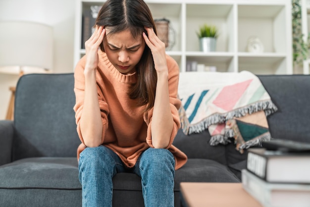 Vermoeide jonge aziatische vrouw die op de bank zit en hoofdpijn heeft vanwege migraine.