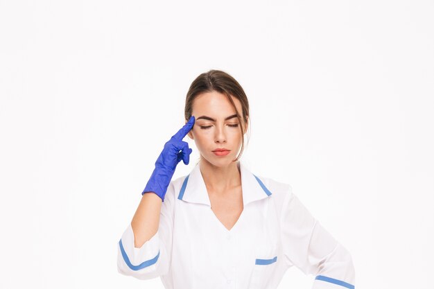 Vermoeide jonge vrouw arts die eenvormige en rubberen handschoenen draagt ?? die zich geïsoleerd over witte muur bevinden