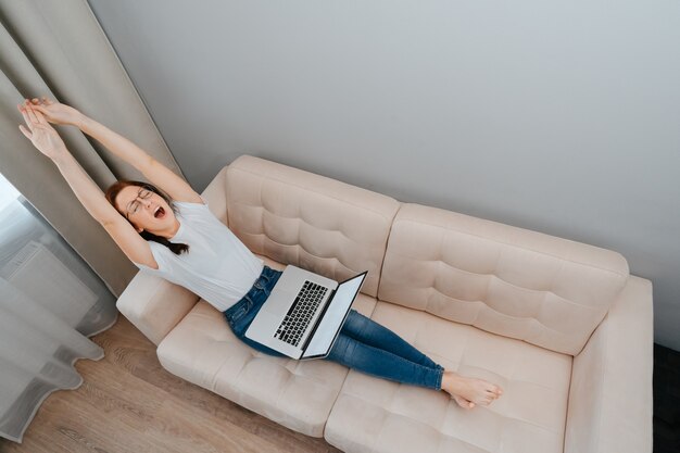 Vermoeide jonge vrouw gaapt terwijl ze op een laptop op een lichte bank thuis zit overwerk op het werk