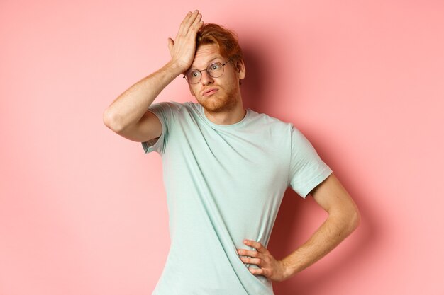 Vermoeide jongeman met rood haar, bril, geïrriteerd en gespannen, facepalm-gebaar maken en uitademen gehinderd, staande over roze achtergrond.