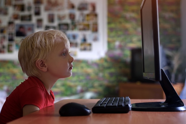 Vermoeide kleine blonde jongen zit zijn neus begraven in een monitor-pc. Internet en kleuter