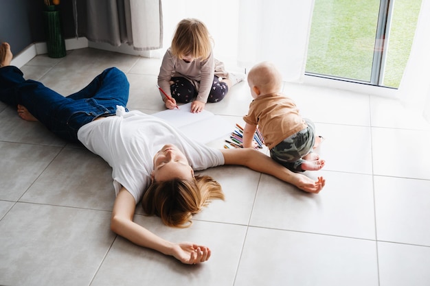 Vermoeide moeder ligt op de vloer terwijl haar kinderen in de buurt tekenen