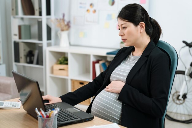 vermoeide overwerkte zwangere vrouw op de werkplek in een helder modern kantoor. uitgeput moederschap lady manager in pak met behulp van laptopcomputer werken. depressieve toekomstige moeder werknemer in pak werk op notebook pc