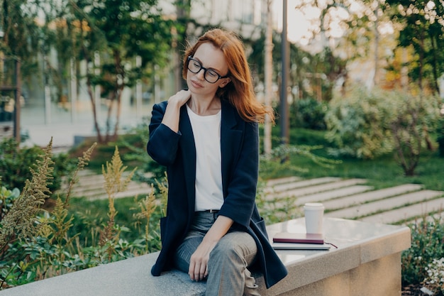 Vermoeide roodharige vrouw neemt rust in park lijdt aan nekpijn zit op bank