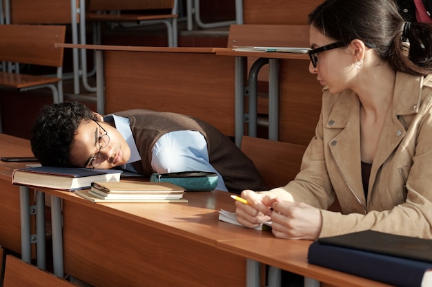 Vermoeide student van gemengd ras die zijn hoofd op het boek houdt terwijl hij naast een vrouwelijke groepsgenoot zit