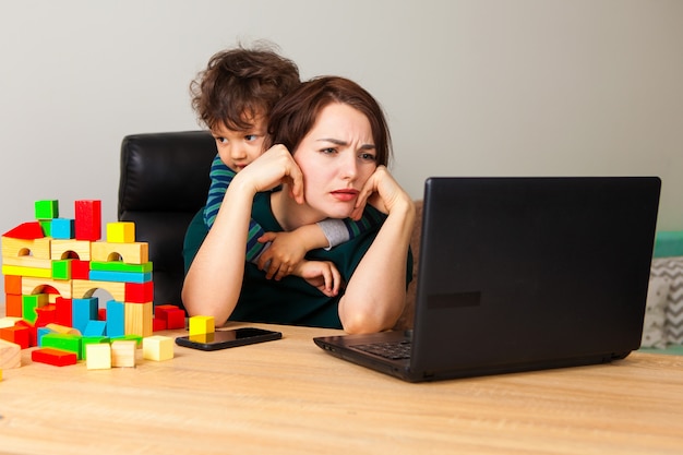 Vermoeide vrouw bij laptop die thuis werkt. Een jongen, een kind bouwde een blokhuis en hing om de nek van zijn moeder en vroeg aandacht voor zichzelf.