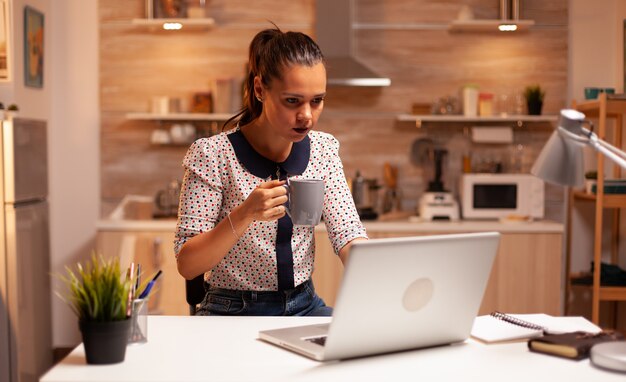 Vermoeide vrouw die 's avonds laat een kopje koffie in de keuken vasthoudt terwijl ze op een laptop werkt. werknemer die om middernacht moderne technologie gebruikt en overuren maakt voor werk, zaken, drukte, carrière, netwerk, levensstijl.