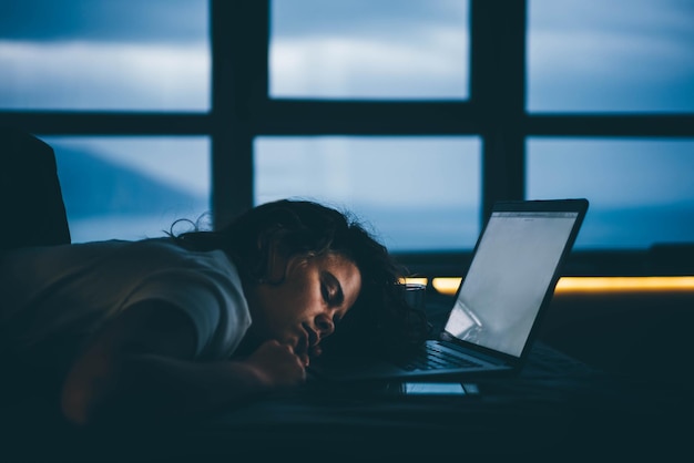 Vermoeide vrouw die 's nachts naast een laptop slaapt.