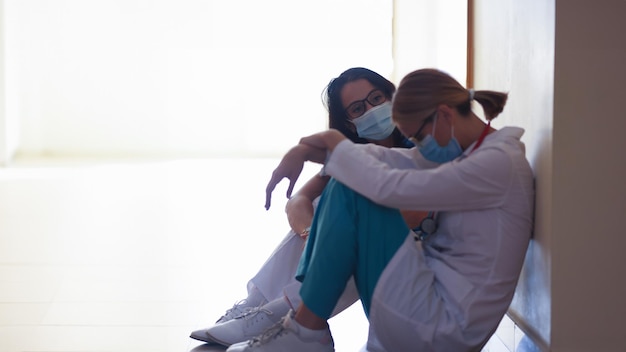 Vermoeide vrouwelijke artsen met beschermende medische maskers zitten in de gang van de kliniek