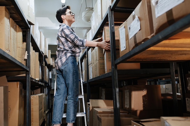 Vermoeide vrouwelijke magazijnmedewerker die op de ladder staat en doos op de plank plaatst