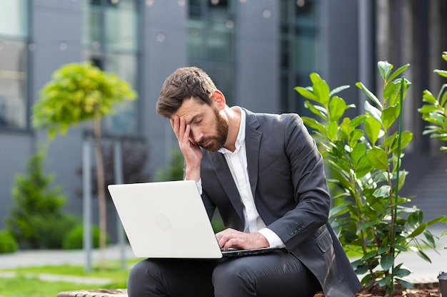 Vermoeide zakenman die op een openbare plaats op een laptopbank werkt, overstuur door depressie en niet tevreden met het resultaat van zijn werk