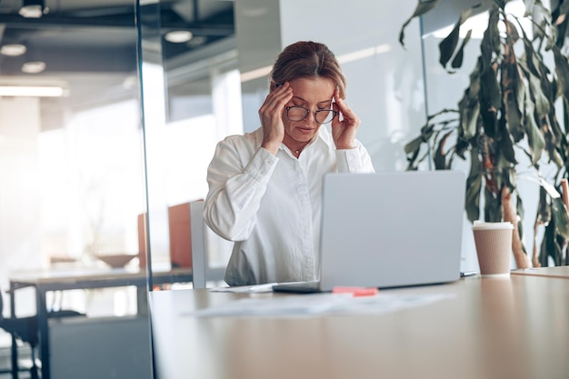 Vermoeide zakenvrouw in glazen met hoofdpijn die op laptop werkt op haar werkplek op modern kantoor