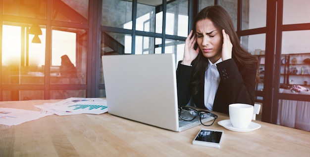Vermoeide zakenvrouw in volledig pak met hoofdpijn terwijl ze op haar laptop in een modern kantoor werkt, houdt haar hoofd vast